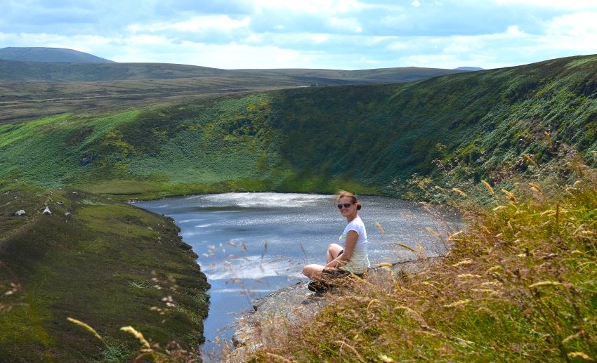 Lato w pełni. Zajmuję miejsce na skale wysoko w Gorach Wicklow w Irlandii, gdzie podziwiam malownicze jezioro rozpościerające się poniżej. Niebo bez chmur rozświetla słońce, a otaczająca przyroda emanuje intensywną zielenią. Moja twarz rozpromieniona uśmiechem odzwierciedla radość z tego niezapomnianego momentu, który chcę zachować na zawsze, przy użyciu aparatu fotograficznego.