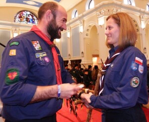 Uroczystość wręczenia nagrody w ramach działalności w Scouts of Ireland. Osoba w niebieskim uniformie Scouts of Ireland uśmiecha się, trzymając w rękach odznakę od Chief 87th group Scouts of Ireland. Oboje wymieniają uścisk dłoni, wyrażając radość i duma z osiągnięcia.