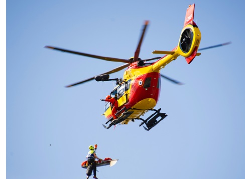 W powietrzu unosi się helikopter ratowniczy, a na linie przymocowanej do niego widnieje ratownik wraz z poszkodowanym w górskiej scenerii. Wizerunek desperackiego wysiłku i nadziei, gdy ratownicy zmagają się z trudno dostępnym terenem, aby uratować życie.